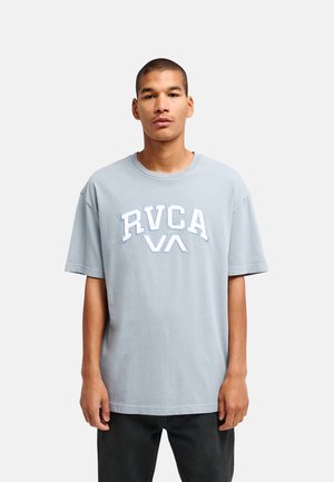 Jeune homme debout face à l'avant portant un t-shirt RVCA gris clair et un pantalon noir sur un fond uni.
