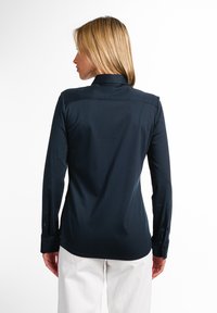 Camicia a maniche lunghe blu navy con colletto, caratterizzata da una texture liscia e polsini con bottoni. La vista posteriore mostra dettagli delle cuciture sulle spalle.