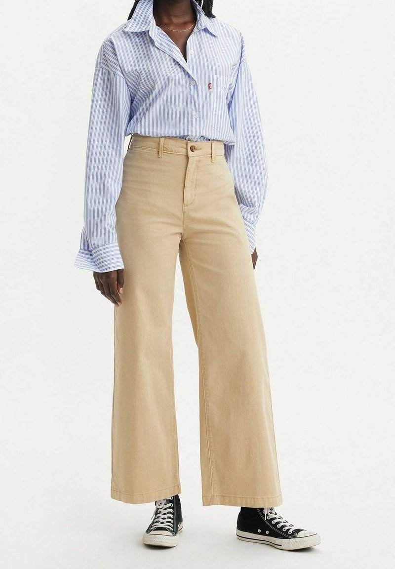 Femme portant une chemise à rayures bleues et blanches avec boutons, un pantalon beige à jambes larges et des baskets montantes noires, debout devant un fond blanc.