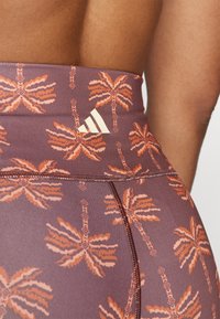 Höga leggings i ett lila tyg med ett palmträdsmönster i orange. Inkluderar en Adidas-logotyp nära midjan och texturerade sömmar.