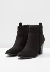 Un par de botas de ante negras hasta el tobillo con tacones de bloque y cremalleras laterales sobre una superficie blanca con un fondo gris claro.