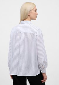 Hvid skjorte med marineblå lodrette striber, lange puffede ærmer, en krave og knapdetaljer ved manchetterne. Materialet virker glat og let.