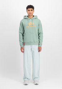 Mentazöld kapucnis pulóver narancssárga logóval, kenguruzsebbel és bordázott mandzsettával. Világoskék, bő szárú farmernadrággal és fehér sportcipővel párosítva.