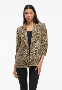 Blazer s uzorkom leoparda u tamnosmeđoj i crnoj tkanini, s oštrim reverima, dva prednja džepa i zavrnutim rukavima, u kombinaciji s crnom majicom.
