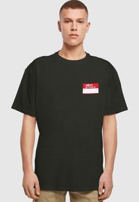 Camiseta de algodón negra con mangas cortas. Presenta un gráfico de etiqueta con nombre en rojo en la parte izquierda del pecho con texto en blanco: "¡HOLA! MI NOMBRE ES."