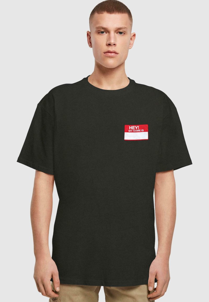 Camiseta de algodón negra con mangas cortas. Presenta un gráfico de etiqueta con nombre en rojo en la parte izquierda del pecho con texto en blanco: "¡HOLA! MI NOMBRE ES."