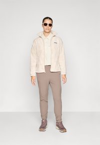 Chaqueta de forro polar de color beige claro con un patrón texturizado, combinada con una camisa crema, pantalones color marrón y zapatos de senderismo morados oscuros. Gafas de sol marrones.