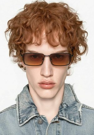 Jeune personne aux cheveux roux bouclés portant des lunettes de soleil rectangulaires teintées marron et une veste en denim claire avec un col effiloché.