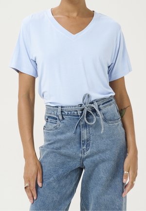 Personne portant un t-shirt bleu clair à col en V et manches courtes, rentré dans un jean bleu taille haute avec une ceinture nouée et un petit tatouage sur le bras gauche.