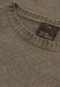 Ruskea neulepusero, jossa on pyöreä ribbikaulus. Kankaan pinta on teksturoitu. Merkkilappu "Oscar Jacobson" on näkyvissä sisäpuolella.