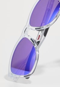 Occhiali da sole con montatura trasparente e lenti viola, caratterizzati da una forma angolare distintiva e sottili accenti neri sulle aste.