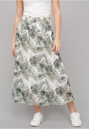Kvinde iført en hvid top og en hvid blomster-midi-nederdel med grønne blade, kombineret med hvide sneakers, stående foran en ensfarvet baggrund.