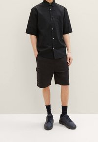 Muž v černé košili s krátkým rukávem na knoflíky, černých cargo šortkách, černých ponožkách a černých teniskách stojící na pozadí v plné béžové barvě.