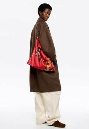 Czerwona torba na ramię z motywem psów, wykonana z gładkiego materiału. Model ma na sobie brązowy płaszcz i kremowe spodnie.