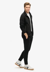 Zwarte zip-up jas met zijzakken, gedragen over een wit T-shirt. Zwarte jogger broek gecombineerd met witte sneakers met zwarte accenten.