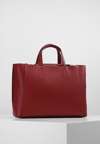 Sac fourre-tout en cuir rouge avec une forme structurée, double anses supérieures et une texture lisse. Présente un design minimaliste avec des accents de couture visibles.