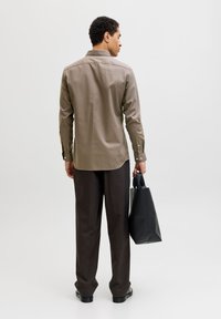 Καφέ πουκάμισο με κουμπιά και καμπύλη ρίγα, συνδυασμένο με σκούρο καφέ παντελόνια. Κρατάει μια μαύρη τσάντα tote. Απλό φόντο.