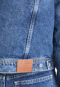 Chaqueta de mezclilla en azul medio con una superficie texturada. Presenta una etiqueta de cuero marrón con el logotipo "TOMMY JEANS" en relieve en la parte trasera de la cintura.