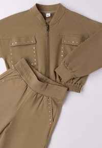 Khaki trainingspak van zachte stof. Voorzien van een rits aan de voorkant, twee borstzakken en studs op de mouwen en broekspijpen. Elastische tailleband.