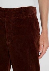 Lähetä kuva henkilöstä, joka pukeutuu tummanruskeisiin corduroy-housuihin, joissa on näkyvä rakenne, ja valkoiseen yläosaan, käsi lepää reiden vierellä.