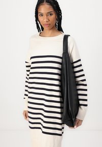 Cream sweater dress with navy horizontal stripes, long sleeves, and a ribbed neckline. Black leather tote bag held at the side.