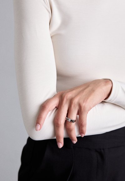 Silberring mit Gravur 'MAMA' und einem klaren Schmuckstein-Akzent, getragen an einer Hand mit heller Haut. Glatte, langärmelige, hellbeige Bluse.