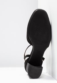 Chaussure pour femme en suede noir avec un petit talon bloc, semelle en caoutchouc visible avec un motif ondulé, positionnée debout contre un mur blanc dans un coin.