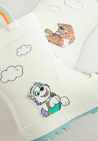 Des bottes de pluie en caoutchouc blanches présentent des personnages d'animaux de bande dessinée et des motifs de nuages. Elles sont dotées d'une semelle texturée bleue et d'une poignée en arc-en-ciel.