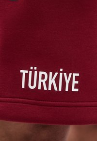 Μαρόν ύφασμα με λευκό κείμενο "TÜRKİYE" εκτυπωμένο κοντά σε ραφή, πιθανώς μέρος ρούχου όπως μανίκι ή μπατζάκι παντελονιού.