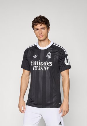 Männliches Model, das ein schwarzes Fußballtrikot von Real Madrid mit dem Logo Emirates Fly Better und weißen Adidas-Shorts auf einem schlichten Hintergrund trägt.