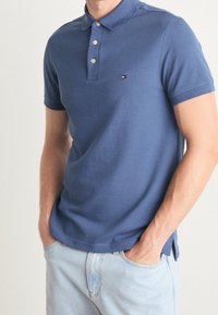 Homme portant un polo bleu à manches courtes avec un petit logo sur la poitrine et un jean bleu clair, mains dans les poches.