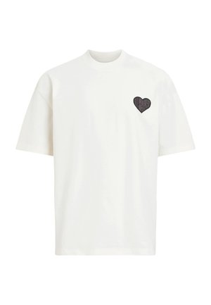 T-shirt bianca a maniche corte in cotone, con una toppa nera a forma di cuore sul lato sinistro del petto con un design testurizzato. Scollo rotondo.