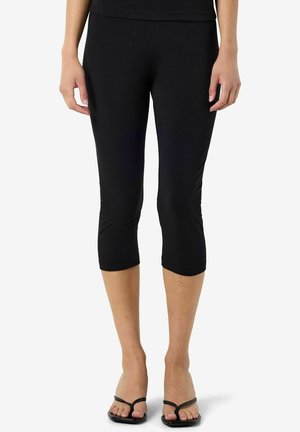 Kvinde iført sorte tætsiddende korte leggings og sorte thongsandaler, stående mod en ensfarvet hvid baggrund.