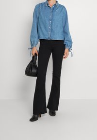 Persona que lleva una camisa de mezclilla azul, pantalones acampanados negros, botas de tacón negras y sostiene un bolso negro frente a un fondo liso.