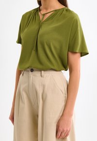 Blouse verte à manches courtes avec une coupe décontractée et un décolleté en V, dotée d'un détail noué au col. Associée à un pantalon taille haute beige clair.