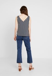 Haut sans manches à rayures bleu marine et blanches avec un dos en V, associé à un jean en denim bleu et des sandales à talons blocs marron. Texture de tissu lisse.