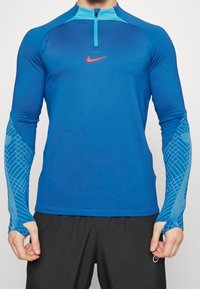 Sininen pitkähihainen urheilupaita, jossa on puolivetoketju kauluksessa, Nike-logo, värilöydöksillä varustettu muotoilu ja kuvioidut yksityiskohdat hihoissa.