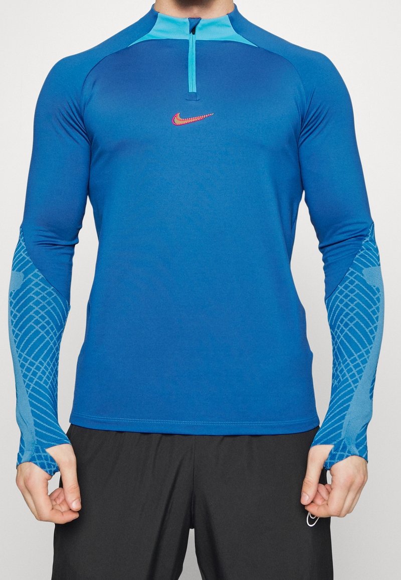 Sininen pitkähihainen urheilupaita, jossa on puolivetoketju kauluksessa, Nike-logo, värilöydöksillä varustettu muotoilu ja kuvioidut yksityiskohdat hihoissa.