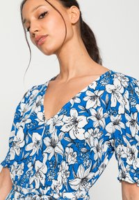 Vestido azul com um padrão floral com flores brancas. Decote em V, mangas curtas com balão e tecido texturizado. Detalhe na cintura com laço.