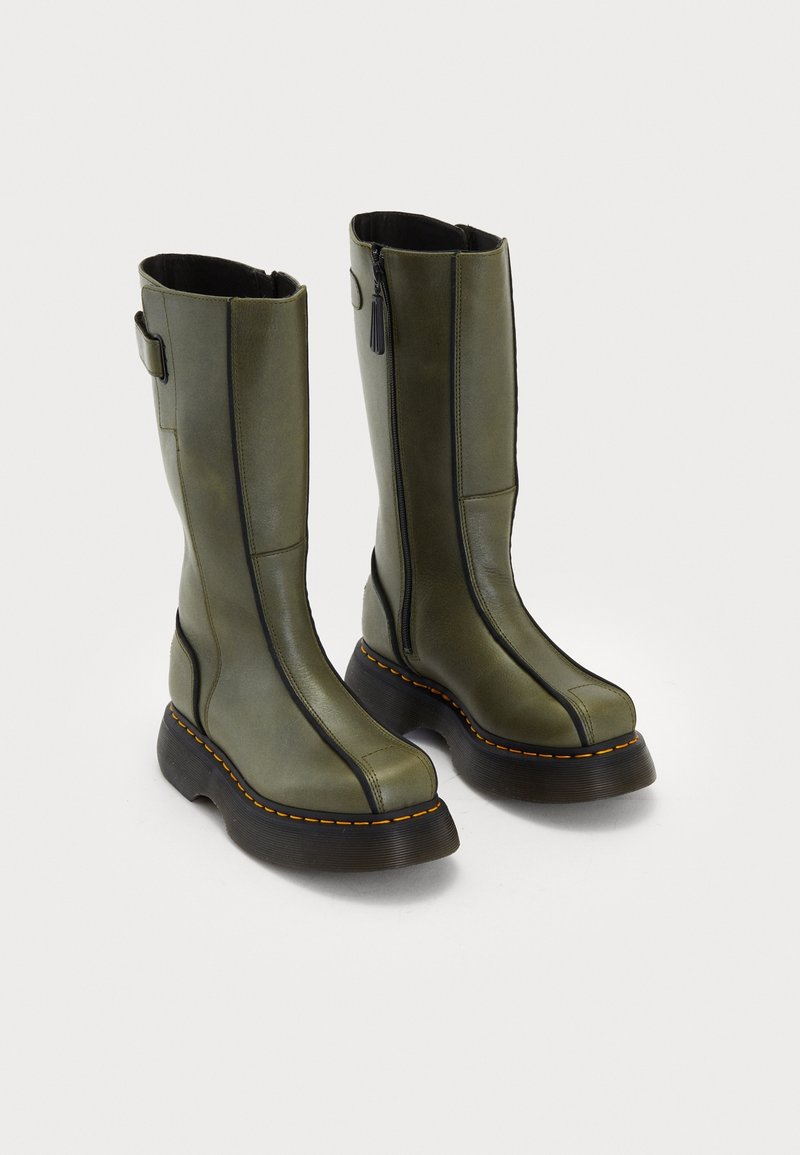 Bottes hautes en cuir vert olive avec semelles épaisses noires, coutures jaunes, fermetures éclair latérales et une bride au talon sur fond blanc.