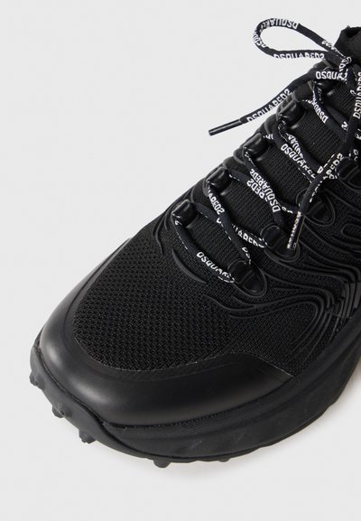 Black athletic shoe featuring mesh upper, rubber outsole, and decorative lacing with repeated brand text. Curved toe and textured details present.
