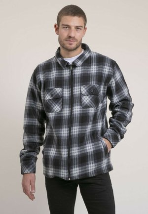 Homme portant une veste zippée à carreaux noirs et gris avec deux poches poitrine à boutons sur le devant, debout avec une main dans la poche.