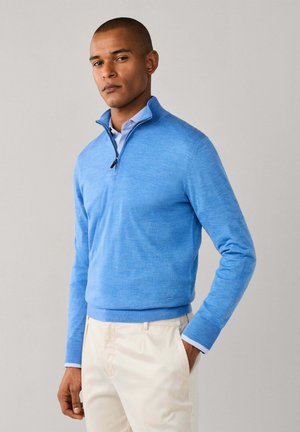 Hombre que lleva un jersey celeste con cremallera hasta el pecho sobre una camisa azul y pantalones beige, de pie con una mano en el bolsillo.