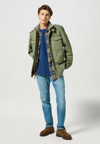 Grön militärjacka över en blå skjorta, matchad med rutig flanell och blå jeans. Bruna skor kompletterar looken.