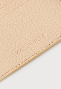 Bežani teksturirani kožni novčanik za kartice s utisnutim logotipom "Coccinelle", s minimalnim šavovima uz rubove radi dodatnih detalja.