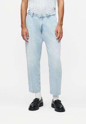 Bărbat purtând blugi largi, cropped, de culoare albastru deschis, șosete albe, mocasini negri și o cămașă cu mâneci scurte cu un model subtil.