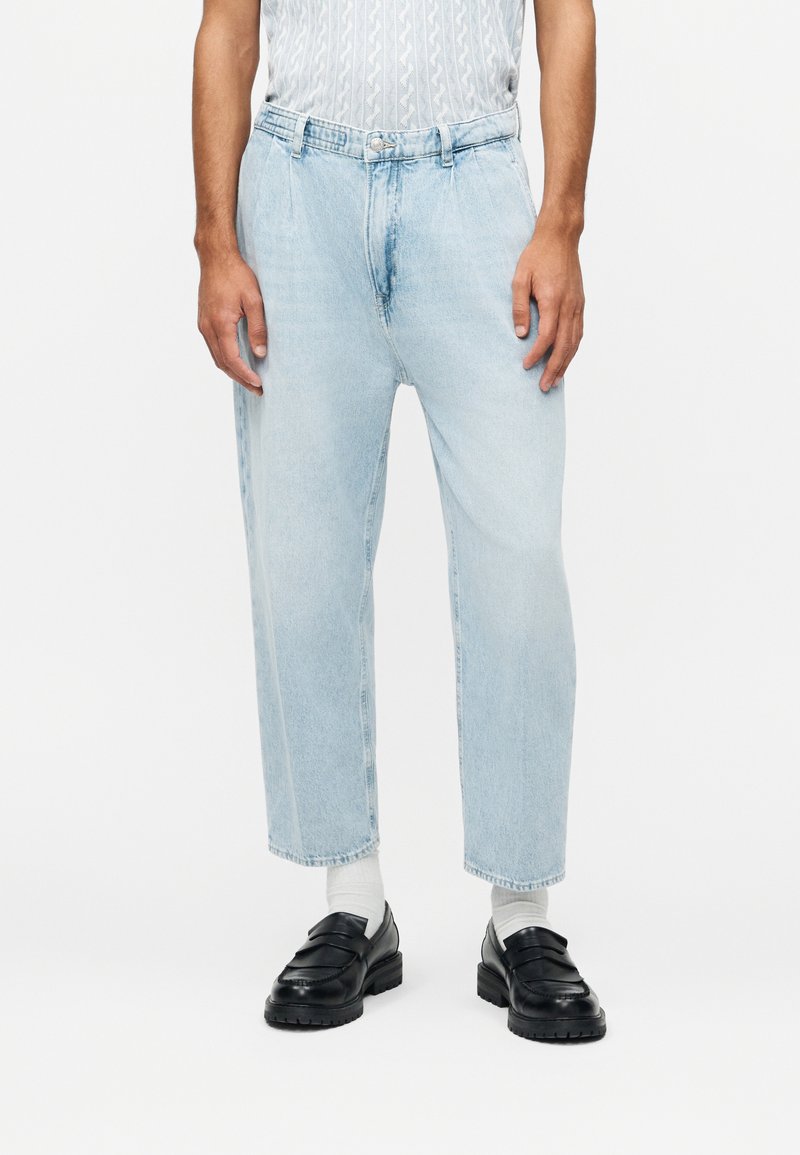 Muž oblečený v svetle modrých zkrácených širokých džínách, bílých ponožkách, černých mokasínách a krátkém rukávu košile s jemným vzorem.