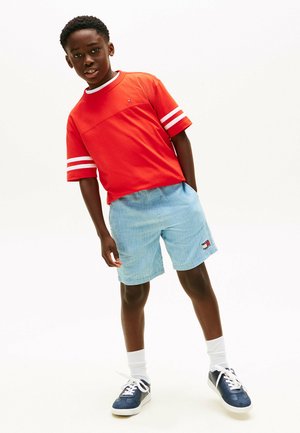 Jongen draagt een rood T-shirt met korte mouwen en witte strepen, lichtblauwe korte broek, witte sokken en marineblauwe sneakers, staat tegen een witte achtergrond.
