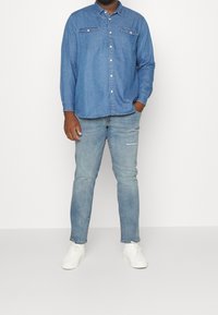 Man wearing light blue denim shirt, light blue ripped jeans, and white sneakers standing against a plain white background.