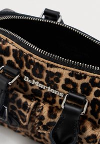 Mala de padrão de leopardo com alças de couro preto, fecho de zíper prateado e detalhe do logo em metal da Dr. Martens no bolso da frente.